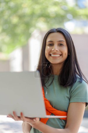 Foto de Retrato de una hermosa mujer india usando una computadora portátil mirando la cámara en la calle de la ciudad. Estudiante, preparándose para el examen, concepto de educación - Imagen libre de derechos