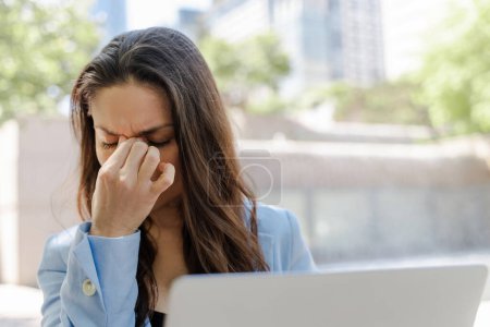 Téléchargez les photos : Femme d'affaires fatiguée à l'aide d'un ordinateur portable frottant les yeux, travaillant en ligne à la rue. Femme stressée ayant mal à la tête lors de la recherche d'informations - en image libre de droit