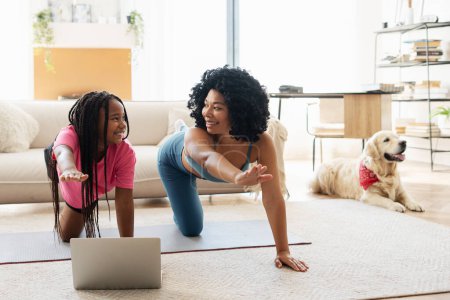 Téléchargez les photos : Souriant afro-américaine petite fille et mère sur tapis de yoga faire des exercices de yoga à la maison. Belle femme et fille faisant de l'exercice ensemble, avec golden retriever. Sport mode de vie sain concept - en image libre de droit