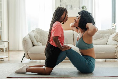 Foto de Atractiva mujer afroamericana haciendo deportes en casa, hija pequeña ayudando, madre haciendo abdominales en el salón. Concepto de transformación corporal, estilo de vida saludable - Imagen libre de derechos