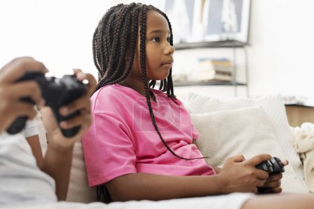 Téléchargez les photos : Belle fille afro-américaine attrayant assis sur le canapé tenant joystick jouer à un jeu vidéo à la maison dans le salon. Enfant sérieux s'amuser en ligne, concept d'enfance - en image libre de droit
