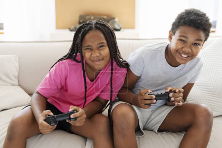 Téléchargez les photos : Portrait de belle soeur afro-américaine positive et frère tenant des joysticks jouant à des jeux vidéo assis sur le canapé dans le salon à la maison. Les enfants positifs s'amusent en ligne. Concept d'enfance - en image libre de droit