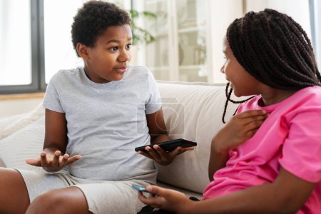 Foto de Retrato de hermosos niños afroamericanos positivos, niños y niñas discutiendo, sentados en el sofá en la sala de estar, en casa. Un chico atractivo se lo explica a la chica. Concepto de relaciones - Imagen libre de derechos