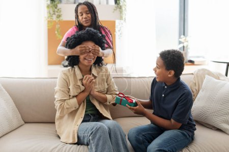Foto de Sonriendo a niños afroamericanos positivos, niños y niñas, cerró los ojos a su madre, dando regalo para el día de la madre, sentado en el sofá en casa en la sala de estar. Concepto de familia feliz - Imagen libre de derechos