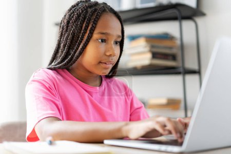Téléchargez les photos : Portrait de fille afro-américaine sérieuse en t-shirt rose tapant sur le clavier, faisant une tâche, apprenant la langue, se préparant à l'examen à la maison. Concept d'éducation en ligne - en image libre de droit