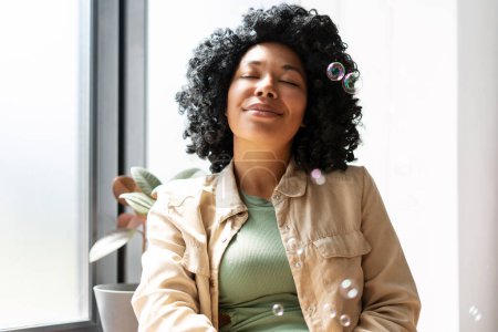Foto de Retrato de una hermosa mujer afroamericana sentada con los ojos cerrados, burbujas de jabón volando en casa en la sala de estar, disfrutando. Concepto de estilo de vida positivo - Imagen libre de derechos