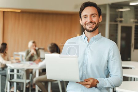 Téléchargez les photos : Portrait d'un gestionnaire hispanique souriant tenant un ordinateur portable debout dans un bureau moderne, vérifiant le courrier, regardant la caméra. Homme d'affaires confiant achats en ligne. Concept de carrière réussie - en image libre de droit