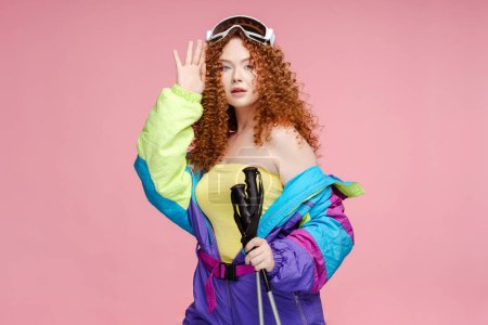 Téléchargez les photos : Femme sexy pensive portant des combinaisons d'hiver élégantes, des lunettes de ski tenant des bâtons de ski, regardant loin isolé sur fond rose. Modèle Fasion, concept hiver - en image libre de droit