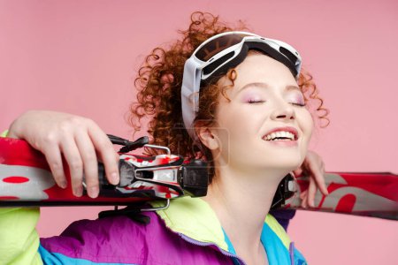 Photo for Attractive smiling woman wearing ski goggles, stylish vintage overalls, holding skis with closed eyes posing isolated on pink background. Advertisement concept - Royalty Free Image