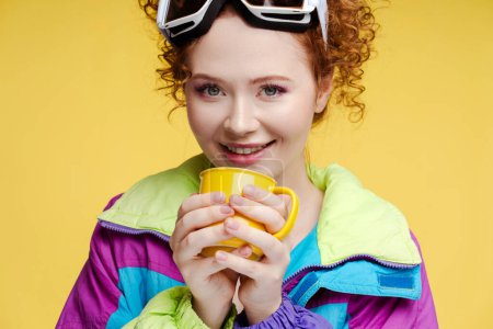 Photo for Closeup positive woman wearing protective ski goggles and stylish vintage overalls, holding cup with drink looking at camera isolated on yellow background. ?oncept of winter, cold - Royalty Free Image
