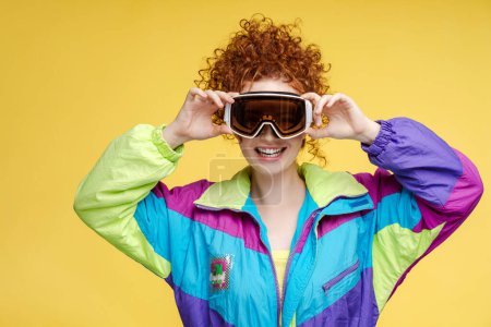 Téléchargez les photos : Portrait d'une belle femme bouclée souriante portant une combinaison de ski, un chapeau, un masque de protection isolé sur fond jaune. Concept de sports d'hiver - en image libre de droit