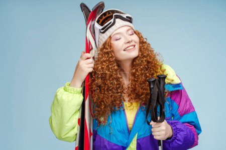 Téléchargez les photos : Portrait de femme séduisante et souriante dans des combinaisons de ski élégantes, tenant des bâtons de ski, des skis, portant des lunettes avec les yeux fermés posant isolés sur fond bleu. Concept de sports d'hiver - en image libre de droit