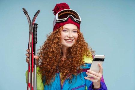Téléchargez les photos : Heureuse femme bouclée dans un chapeau avec des lunettes de ski de protection tenant matériel de ski à l'aide de téléphone mobile, achats en ligne, la commande en regardant la caméra isolée sur fond bleu. Concept Internet - en image libre de droit
