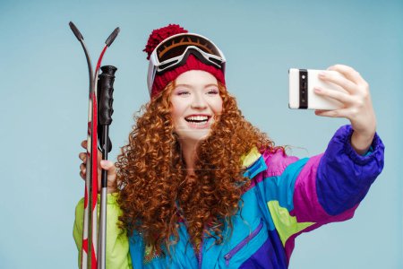 Photo for Smiling curly woman in hat with protective ski goggles holding ski gear using mobile phone, taking selfie, recording video isolated on blue background. Technology concept - Royalty Free Image