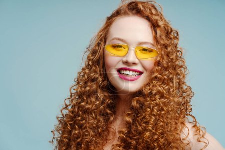 Photo for Portrait of beautiful smiling curly redhead woman wearing sunglasses looking at camera isolated on blue background. Fashion concept, advertisement - Royalty Free Image