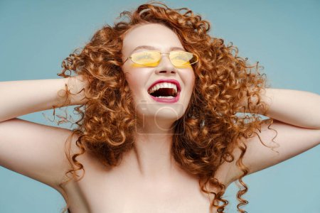 Foto de Retrato de mujer pelirroja rizada emocionada sonriente con gafas de sol mirando hacia otro lado aislada sobre fondo azul. Concepto de moda, publicidad - Imagen libre de derechos