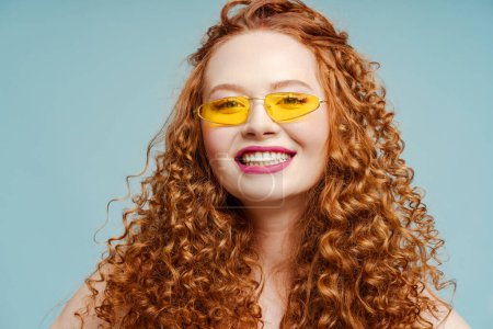 Foto de Atractiva mujer pelirroja rizada sonriente con gafas de sol mirando a la cámara aislada sobre fondo azul. Concepto de cuidado dental - Imagen libre de derechos