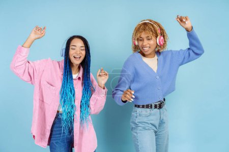 Photo for Portrait of happy beautiful girls in colorful casual clothes in wireless headphones listening to music, having fun, dancing isolated on blue background. Hobby concept - Royalty Free Image