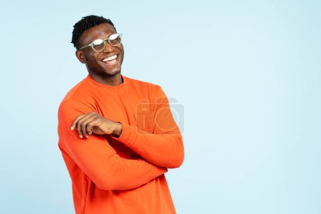 Téléchargez les photos : Portrait d'un homme souriant et attrayant afro-américain portant un t-shirt orange décontracté, des lunettes élégantes isolées sur fond bleu. Concept de shopping, publicité - en image libre de droit