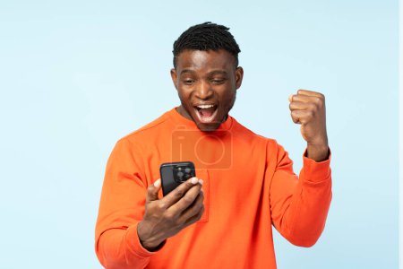 Téléchargez les photos : Jeune homme afro-américain ravi, joueur tenant téléphone mobile paris sportifs, gagner succès de célébration de l'argent isolé sur fond. Heureux gars faisant du shopping en ligne avec des ventes. Innovation technologique - en image libre de droit