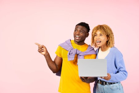 Téléchargez les photos : Jeunes amis souriants tenant un ordinateur portable, magasinant en ligne pointant du doigt sur l'espace de copie, isolé sur fond rose. Étudiants heureux regardant ailleurs, concept d'éducation - en image libre de droit