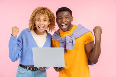 Téléchargez les photos : Couple d'amis souriants tenant ordinateur portable célébration succès isolé sur fond rose. Heureux homme et femme afro-américain élégant faisant du shopping en ligne en regardant la caméra. Technologie - en image libre de droit