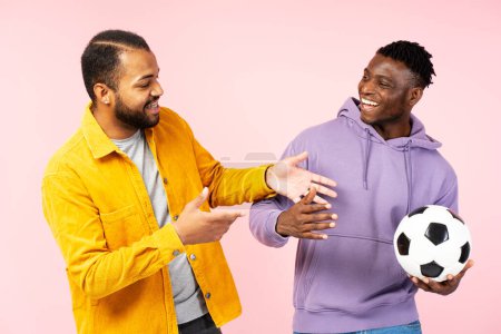 Téléchargez les photos : Heureux souriants amis afro-américains, joueurs de football tenant ballon de football, parler portant tenue élégante isolé sur fond rose. Concept de sport, football - en image libre de droit