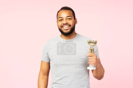Téléchargez les photos : Overjoy souriant homme afro-américain portant t-shirt occasionnel gris tenant coupe trophée isolé sur fond rose. Homme positif portant des accolades regardant la caméra. Concept de victoire - en image libre de droit