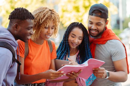 Téléchargez les photos : Groupe de jeunes étudiants afro-américains souriants lisant le livre, se préparant à l'examen, debout près du campus universitaire dans la rue. Des jeunes heureux se rencontrent à l'extérieur - en image libre de droit