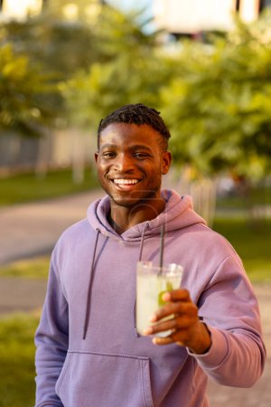 Foto de Guapo sonriente elegante afroamericano joven sosteniendo limonada, beber, mirando a la cámara en la calle, al aire libre - Imagen libre de derechos