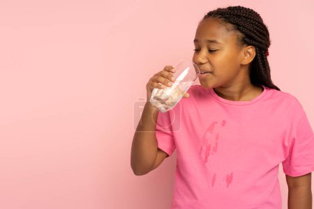 Photo for Beautiful positive African girl with stylish hairstyle drinking water from glass standing isolated on pink background, copy space. Cute female spilled water on her t shirt - Royalty Free Image