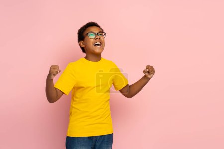 Photo for Portrait of excited smiling Nigerian boy wearing glasses and stylish yellow t shirt and jeans making winner gesture, looking away isolated on pink background, copy space. Concept of victory - Royalty Free Image