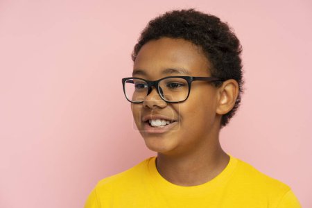 Foto de Retrato de niño de escuela afroamericano guapo sonriente con gafas con estilo y camiseta amarilla mirando hacia otro lado aislado sobre fondo rosa. Concepto educativo - Imagen libre de derechos