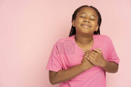 Téléchargez les photos : Portrait de mignonne belle petite afro-américaine tenant la main sur le cœur avec les yeux fermés rêvant, vêtue d'un t-shirt rose décontracté isolé sur fond rose, espace de copie - en image libre de droit