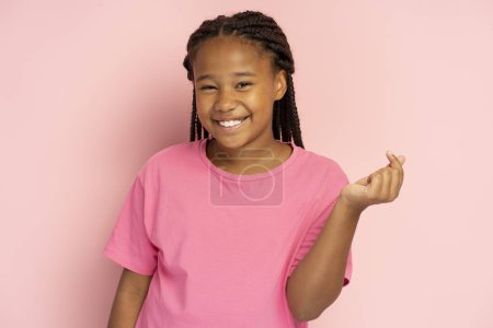 Porträt eines lächelnden afroamerikanischen Mädchens in rosa T-Shirt, das gestikulierend Herz von Fingern zeigt, k Popkultur, isoliert auf rosa Hintergrund, Attrappe. Modernes Kind mit zahmem Lächeln, das in die Kamera blickt