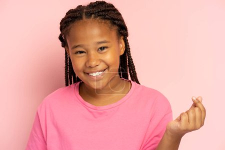 Schöne kleine afroamerikanische Mädchen gestikulierend, die Herz von den Fingern zeigt, k Popkultur, isoliert auf rosa Hintergrund, Attrappe. Glückliches modernes Kind mit zahmem Lächeln, das in die Kamera blickt