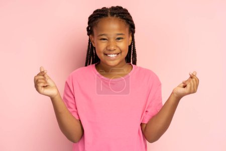 Fille africaine positive en t-shirt rose geste montrant le c?ur des doigts, k pop culture, isolé sur fond rose, maquette. Jeune enfant moderne avec le sourire de dent regardant la caméra