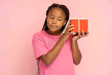 Téléchargez les photos : Portrait de belle petite fille afro-américaine tenant boîte cadeau rouge isolé sur fond rose. Enfant mignon a reçu un cadeau pour le Nouvel An. Concept de célébration - en image libre de droit