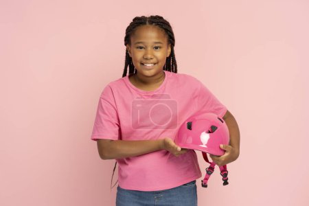 Foto de Sonriente hermosa chica africana sosteniendo casco rosa con ropa casual con estilo, peinado elegante, rastas de pie aislado sobre fondo rosa. Atractivo niño posando mirando a la cámara - Imagen libre de derechos