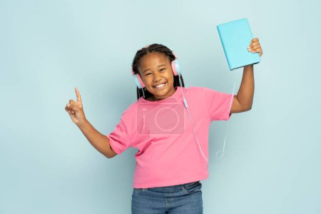 Téléchargez les photos : Portrait de fille africaine souriante avec une coiffure élégante portant t-shirt décontracté écoutant de la musique dans les écouteurs tenant tablette numérique regardant loin isolé sur fond bleu, espace de copie - en image libre de droit
