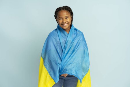 Foto de Sonriente chica nigeriana positiva sosteniendo bandera ucraniana envuelta en ella mirando a la cámara aislada sobre fondo azul, espacio de copia. Concepto de símbolo nacional, patriotismo - Imagen libre de derechos