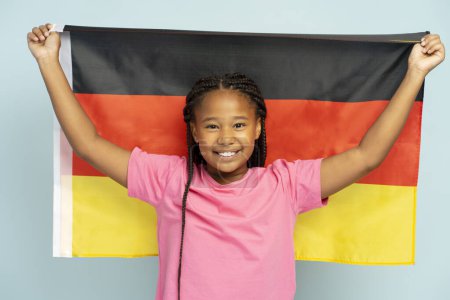 Téléchargez les photos : Portrait d'une jolie Africaine souriante tenant le drapeau allemand portant un t-shirt rose décontracté isolé sur fond bleu. ? Acceptation du symbole national, soutien, fête de l'indépendance - en image libre de droit