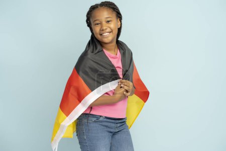 Photo for Portrait of positive Nigerian girl holding German flag wrapped in it looking at camera isolated on blue background, copy space. Concept of national symbol, patriotism - Royalty Free Image
