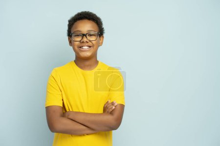 Foto de Retrato de niño afroamericano guapo sonriente en camiseta casual amarilla con gafas con brazos cruzados de pie aislado sobre fondo azul, espacio para copiar. Concepto educativo - Imagen libre de derechos