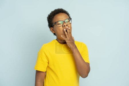 Téléchargez les photos : Portrait d'un adolescent afro-américain fatigué portant un t-shirt jaune élégant et des lunettes bâillant, se réveiller le matin, isolé sur fond bleu - en image libre de droit