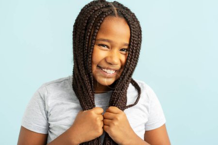 Foto de Retrato de sonrisa positiva hermosa chica afroamericana con elegantes coletas, peinado sosteniendo el pelo mirando en el espejo aislado sobre fondo azul. Concepto de cuidado del cabello - Imagen libre de derechos