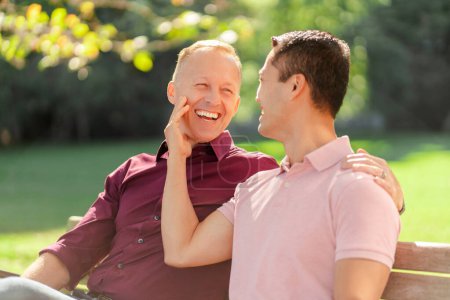 Téléchargez les photos : Un beau couple gay souriant, deux hommes parlant, se regardant s'embrasser assis sur le banc du parc. Lgbt concept, rencontres, relation - en image libre de droit