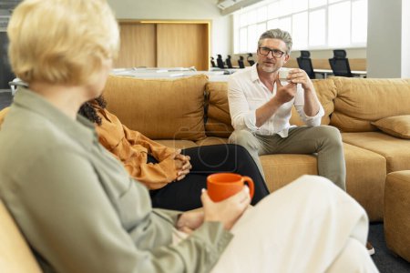 Foto de Retrato de empresarios exitosos sentados en un cómodo sofá sosteniendo la taza bebiendo bebida disfrutando de una pausa para tomar café en la oficina moderna. Concepto empresarial - Imagen libre de derechos