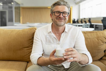 Foto de Retrato de un hombre de negocios de pelo gris sonriente sentado en un cómodo sofá sosteniendo la taza con los ojos cerrados bebiendo bebida disfrutando de un descanso de café en la oficina moderna, espacio para copiar - Imagen libre de derechos