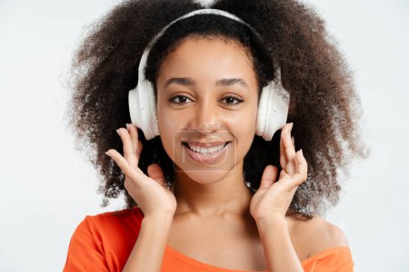 Foto de Retrato de una mujer afroamericana sonriente y amigable con el pelo rizado escuchando música en auriculares mirando a la cámara, de cerca. Concepto de tecnología, diversión - Imagen libre de derechos